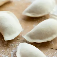 Surowe pierogi na drewnianej desce, posypane mąką. Idealne ciasto na pierogi bez jajka, gotowe do nadziewania.