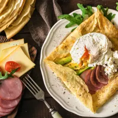 Stos **crepes** z jajkiem w koszulce, awokado, szynką i serem. Obok stos naleśników, pomidorki i rukola.