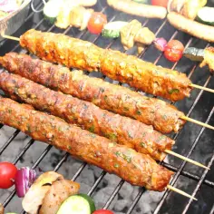 Szaszłyki z mięsa mielonego, pachnące ziołami, pieką się na grillu obok warzyw.