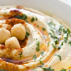 Kremowy hummus z ciecierzycy, polany oliwą, posypany papryką i natką pietruszki. Na wierzchu trzy całe ziarna ciecierzycy.