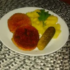 Dwa złociste kotlety z mortadeli, puree ziemniaczane z natką pietruszki i ogórek kiszony na białym talerzu.