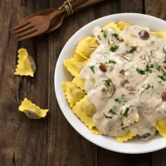 Ravioli z grzybami w kremowym sosie śmietanowym, posypane natką pietruszki. Pyszny obiad na drewnianym stole.