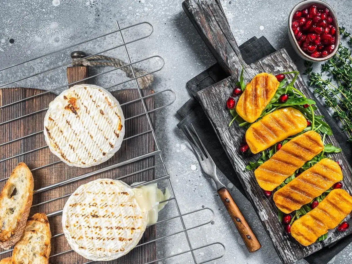 Dwa krążki serów na grilla, jeden lekko roztopiony, obok grzanki. Na desce obok grillowane kawałki dyni z rukolą i granatem.