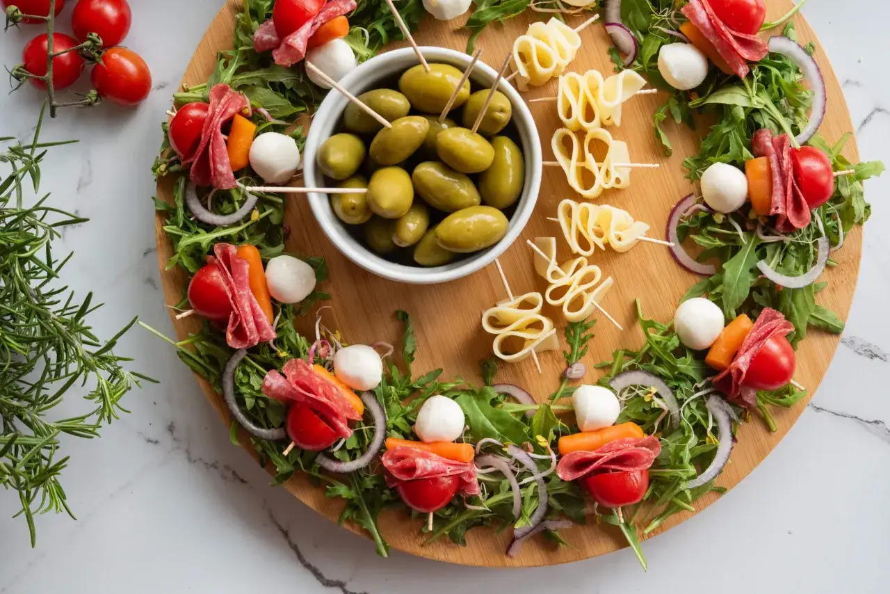Okrągła deska z apetycznymi, słonymi przekąskami: szaszłyki z mozzarellą, salami, pomidorkami i marchewką, oliwki, serowe zawijasy.