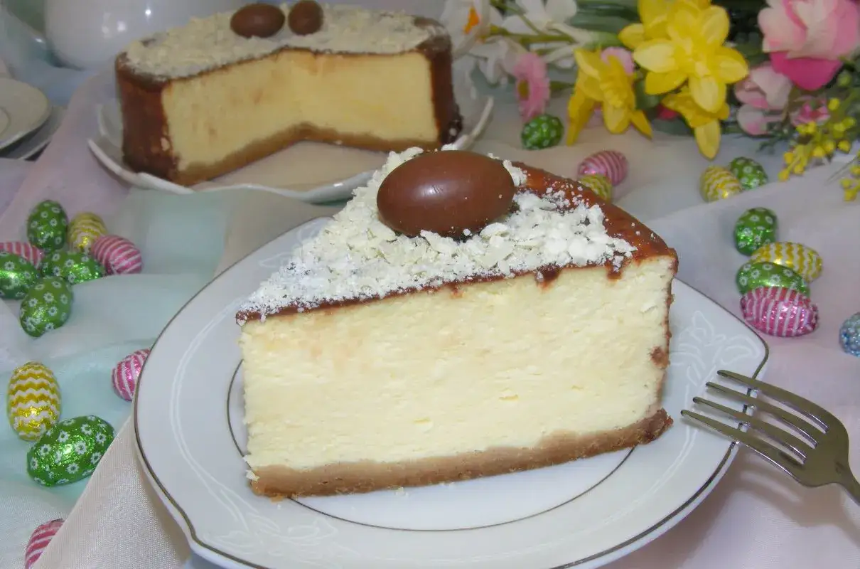 Prosty sernik z serka mascarpone, udekorowany czekoladowymi jajeczkami i wiórkami białej czekolady, na tle wielkanocnych ozdób.