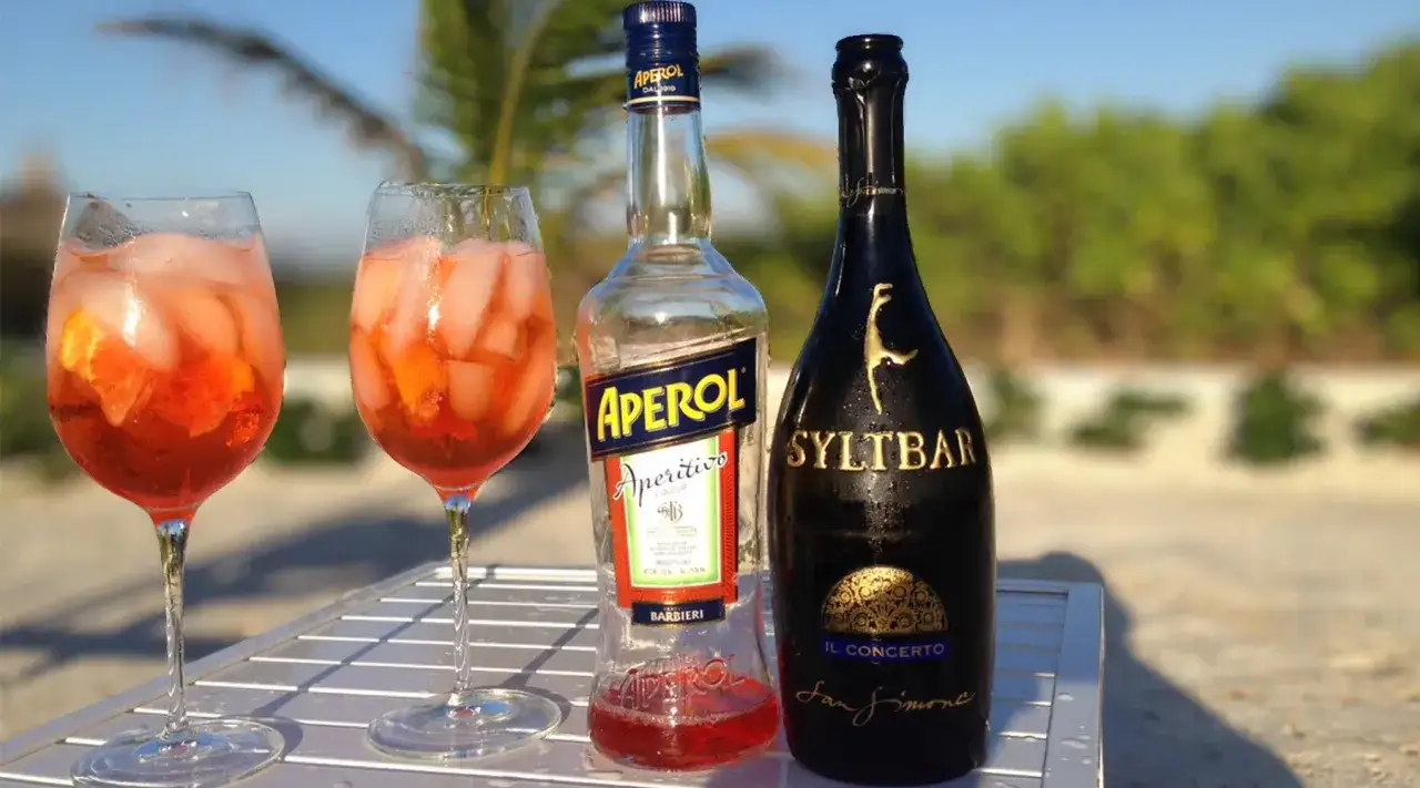 Dwa drinki Aperol Spritz z lodem i pomarańczą, butelka Aperolu i butelka Syltbar. Idealne na aperitivo, zastanawiasz się jakie prosecco do Aperola?