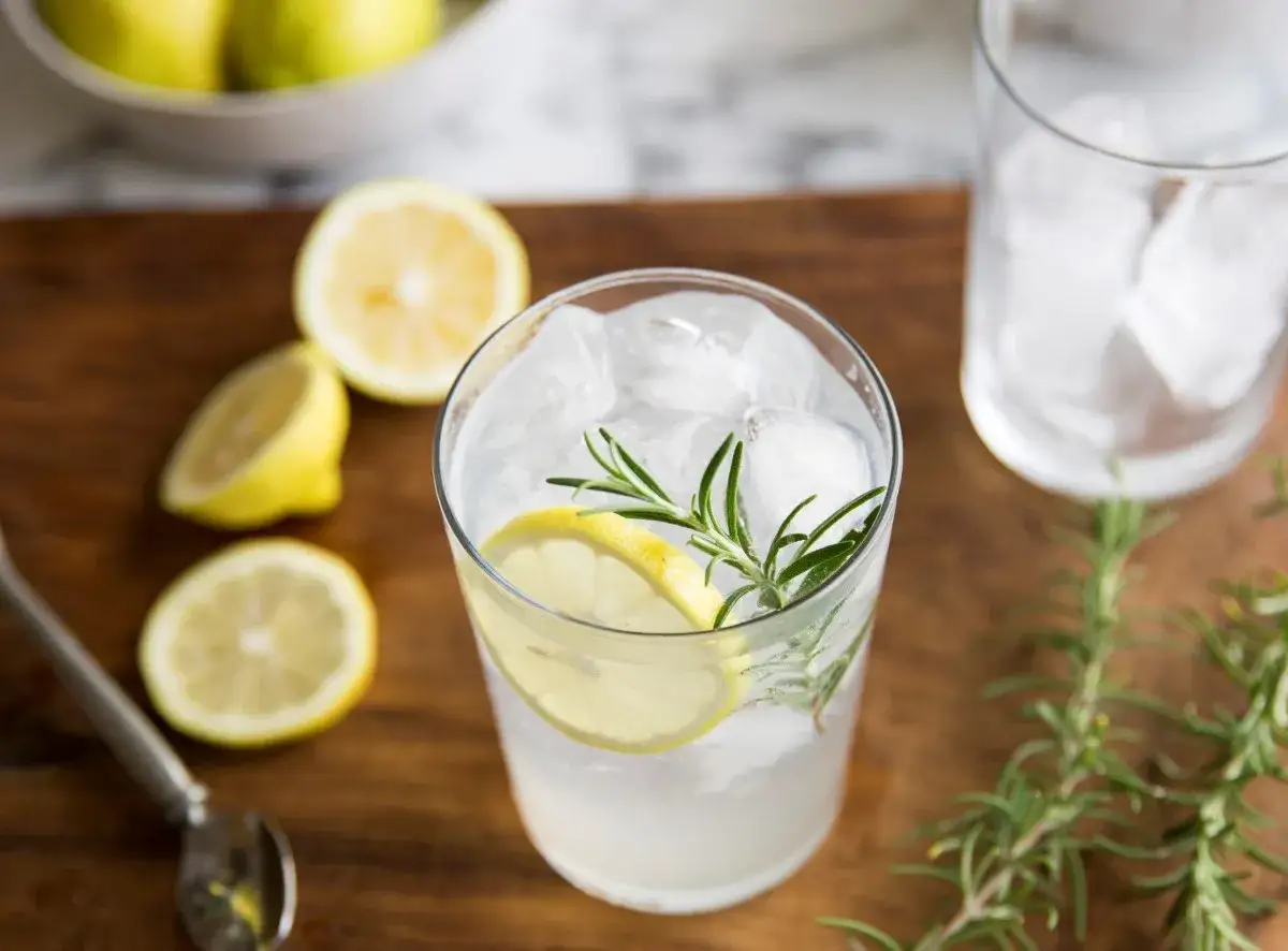 Orzeźwiający gin z tonikiem, idealne proporcje z cytryną i rozmarynem. Obok szklanka z lodem i cytryny.