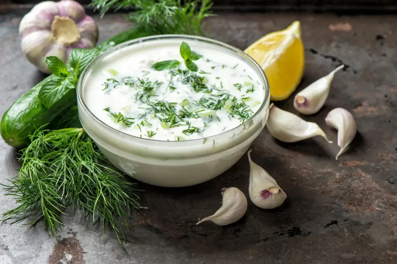 Kremowy sos aioli z czosnkiem, ogórkiem i ziołami, obok cytryny i świeżych składników.