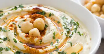 Kremowy hummus z ciecierzycy, polany oliwą, posypany papryką i natką pietruszki. Na wierzchu trzy całe ziarna ciecierzycy.