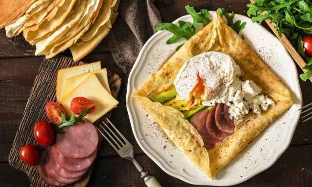 Stos **crepes** z jajkiem w koszulce, awokado, szynką i serem. Obok stos naleśników, pomidorki i rukola.